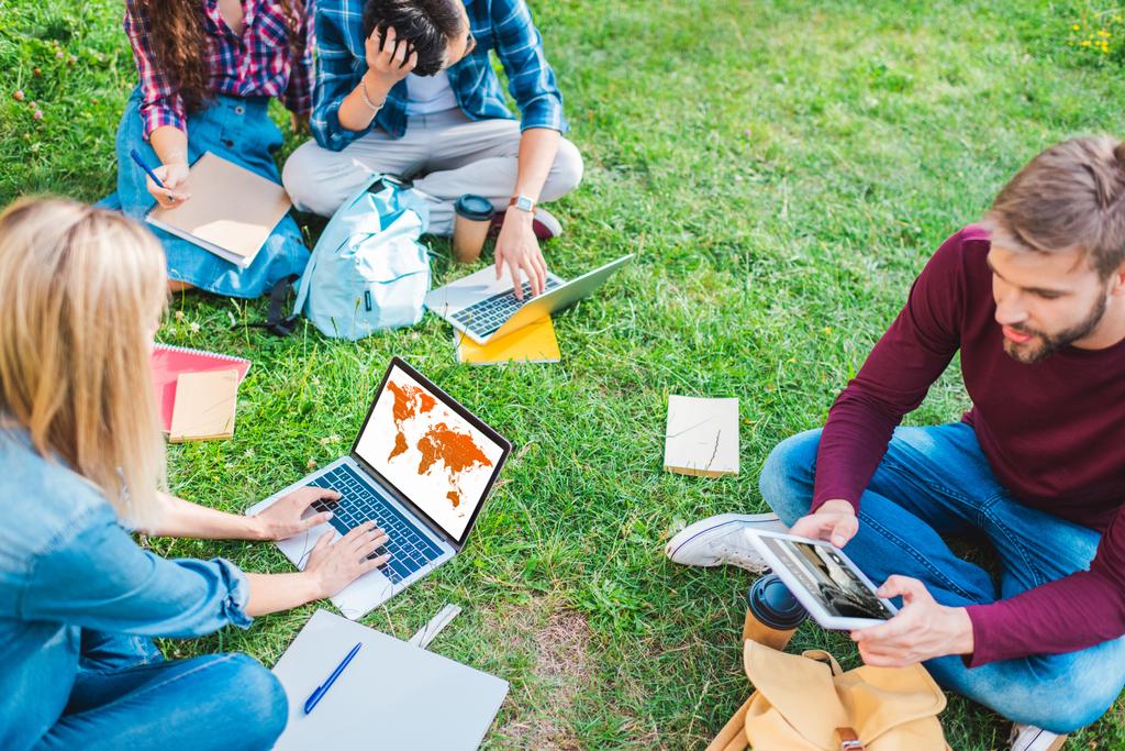 ノートブックおよび公園の緑の草の上に座ってデジタル デバイスと多民族の学生の部分的なビュー - 写真・画像