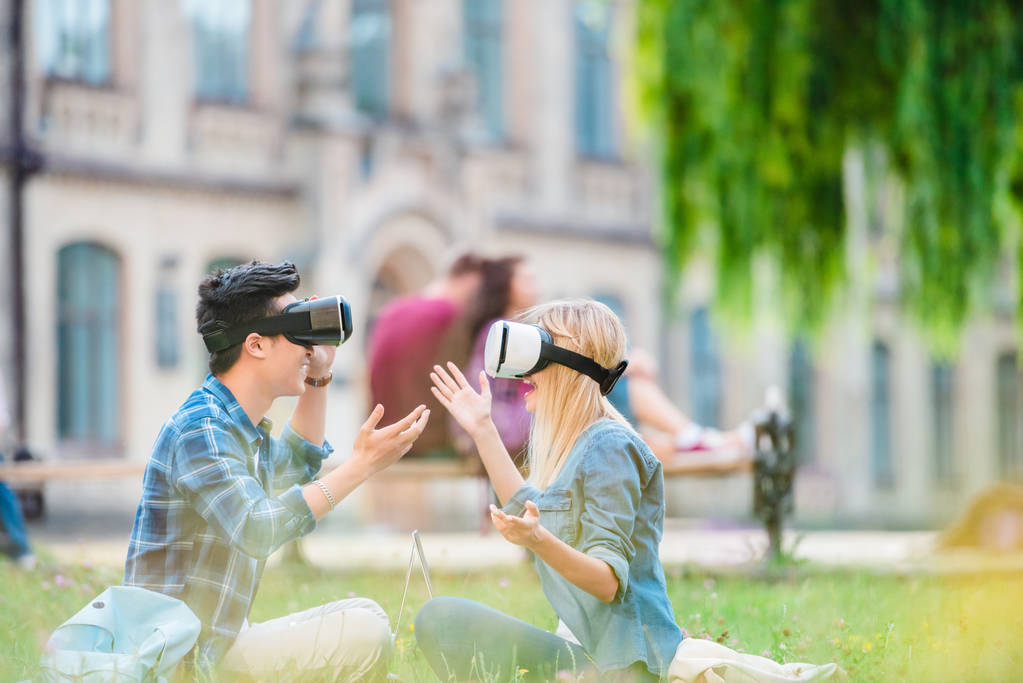 side view of multicultural students in virtual reality headsets with laptops sitting on green grass in park - Photo, Image