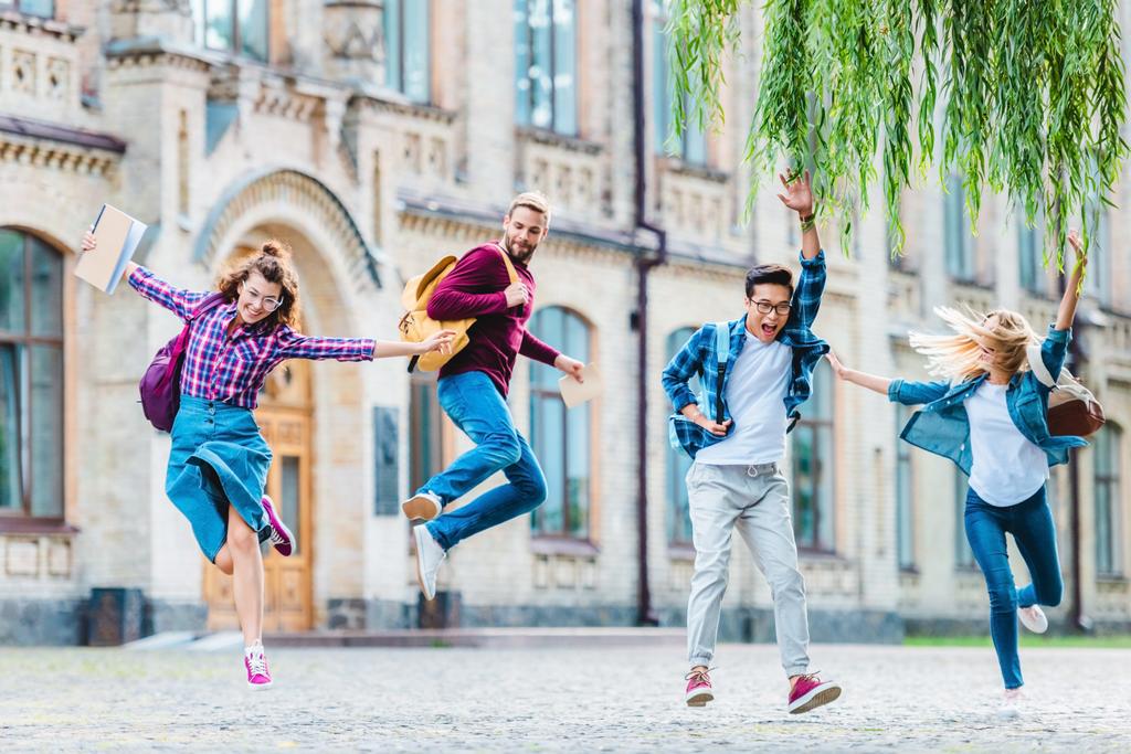 happy multicultural students with backpacks jumping on street with university on background - Photo, Image