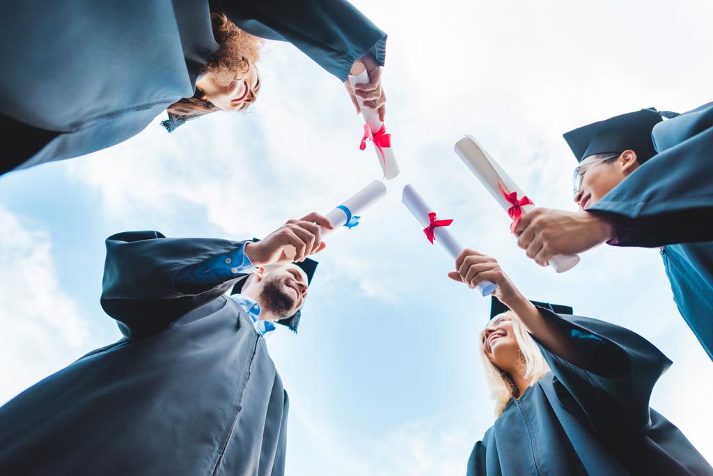 pohled zdola mnohonárodnostní absolventů s diplomy v ruce a modré obloze na pozadí - Fotografie, Obrázek