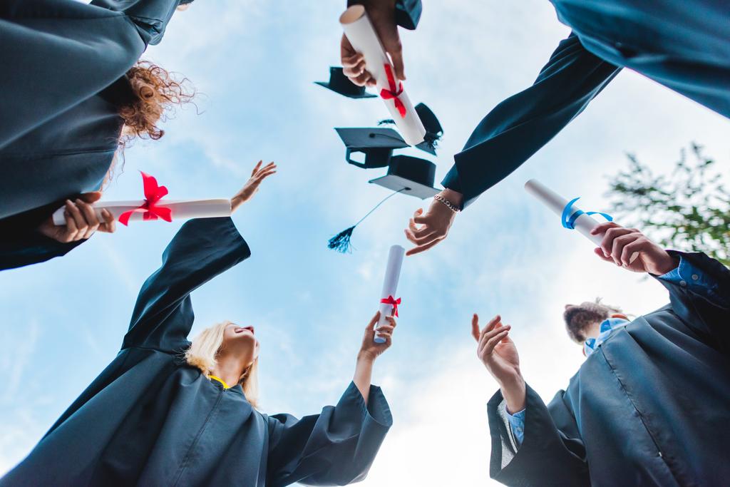 Blick von unten auf glückliche multikulturelle Absolventen mit Diplomen, die mit blauem Himmel auf dem Hintergrund Mützen hochwerfen - Foto, Bild