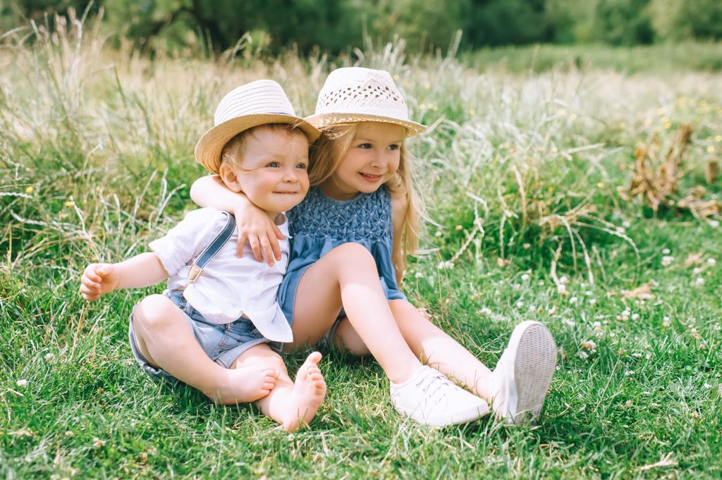 緑の野原に座っている麦わら帽子でかわいいスタイリッシュな子供たち  - 写真・画像