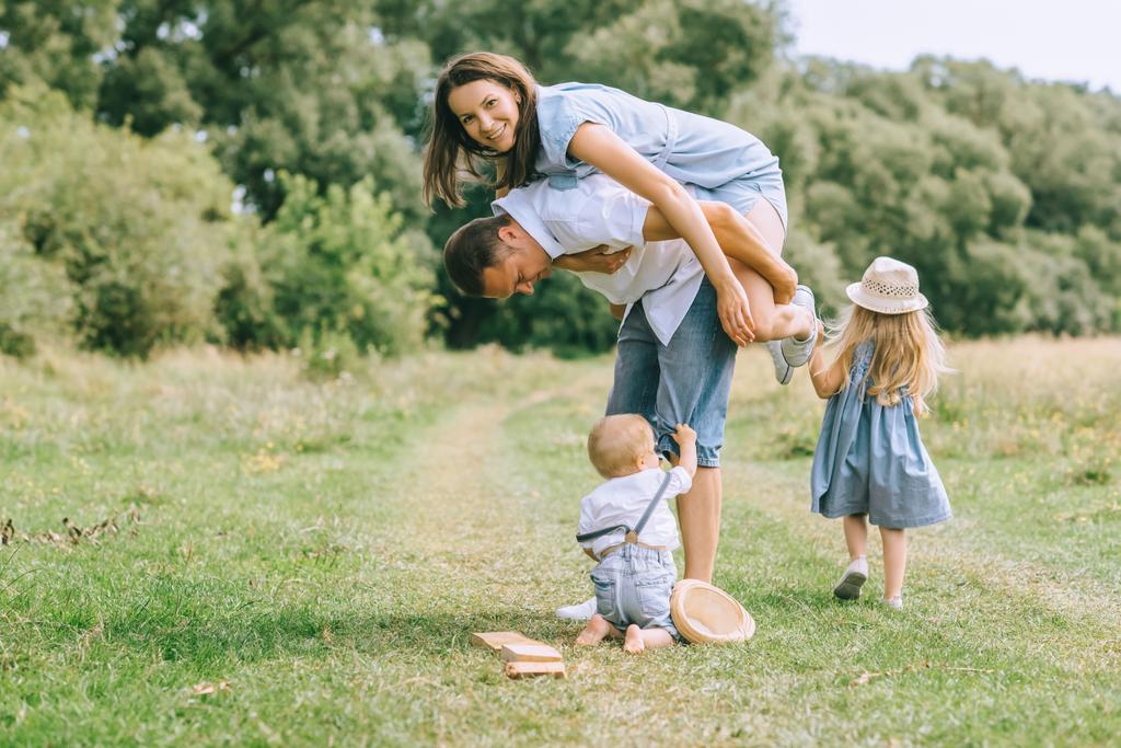 ευτυχισμένη οικογένεια με δύο παιδιά που αφιερώνουν χρόνο στο πεδίο καλοκαίρι, πατέρας piggybacking μαμά - Φωτογραφία, εικόνα