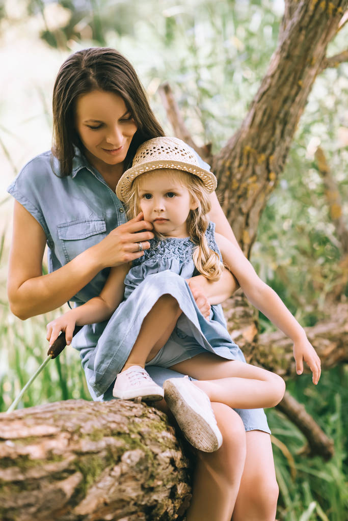 happy mother and adorable blonde daughter sitting on tree trunk - Photo, Image