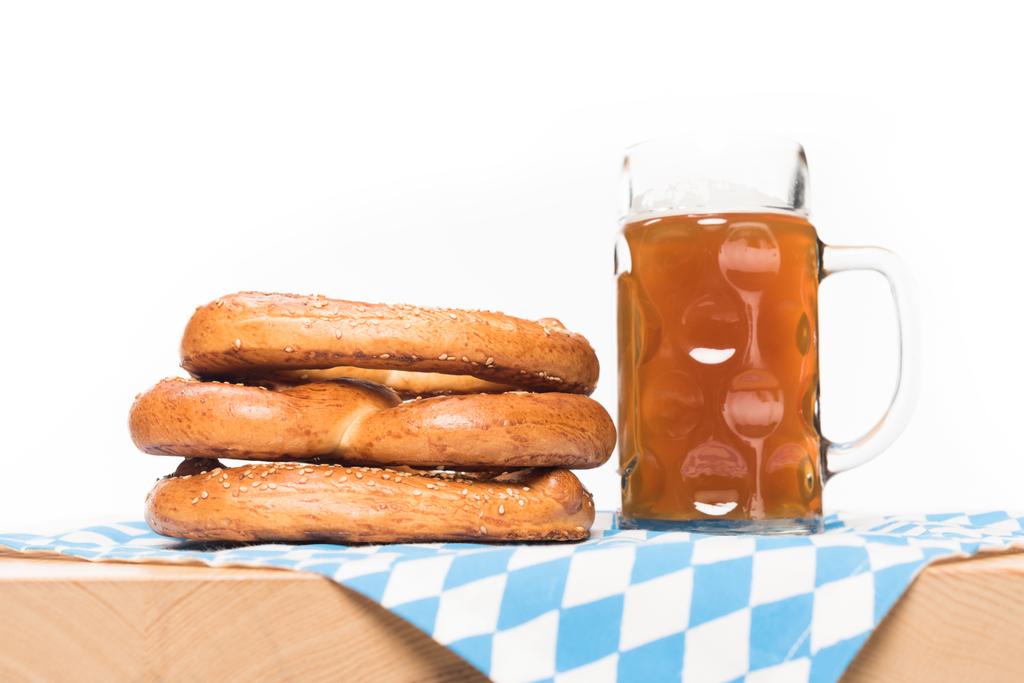 επιλεκτική εστίαση του κούπα της μπύρας και pretzels στο τραπέζι με τραπεζομάντιλο σε λευκό φόντο  - Φωτογραφία, εικόνα