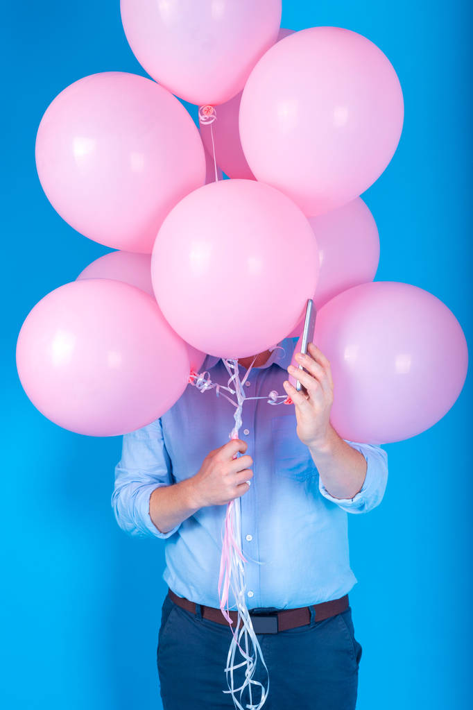 Młody przystojny mężczyzna w niebieską koszulę, trzymając różowy balony i smartphone na niebieskim tle, wolne miejsca na tekst. Wakacje, urodziny, pojęcie dostawy - Zdjęcie, obraz