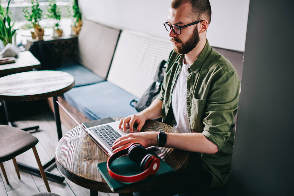 Серьезный опытный кавказский программист в очках сидит в кафе с чердаком и работает удаленно, молодой вдумчивый человек смотрит вебинар за ноутбуком и печатает идеи для курсового текста
 - Фото, изображение