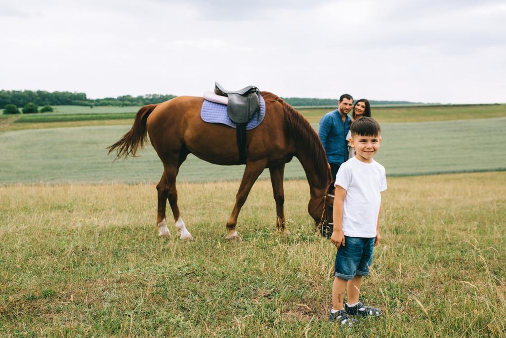ouders staan in de buurt van paard, glimlachend zoon kijken camera op veld - Foto, afbeelding
