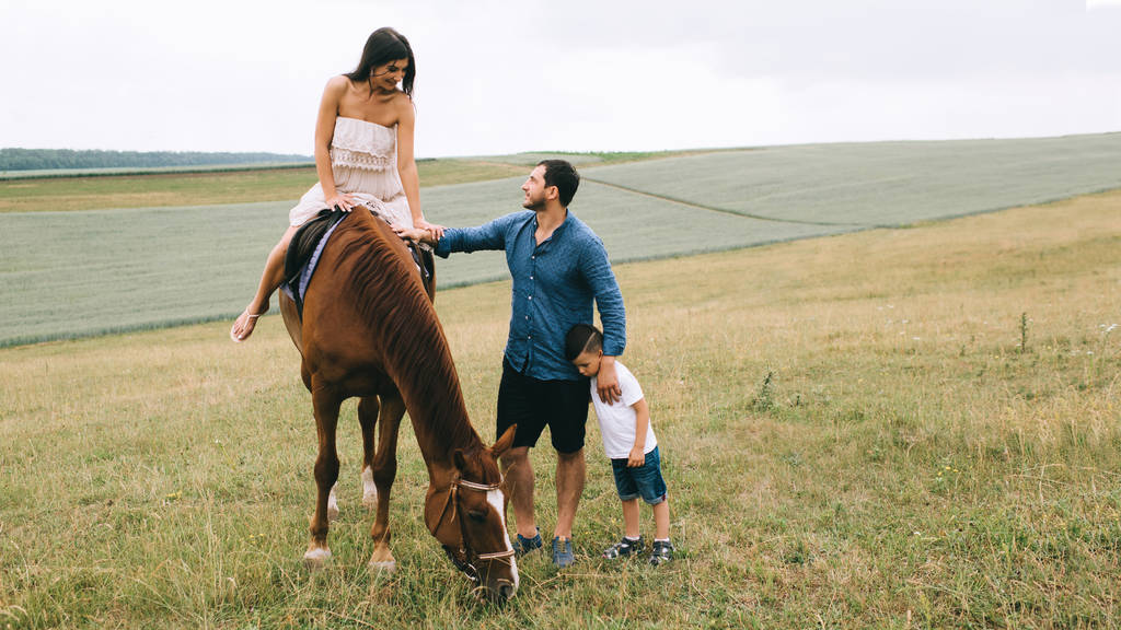 Mutter reitet Pferd, Vater und Sohn stehen auf Feld - Foto, Bild