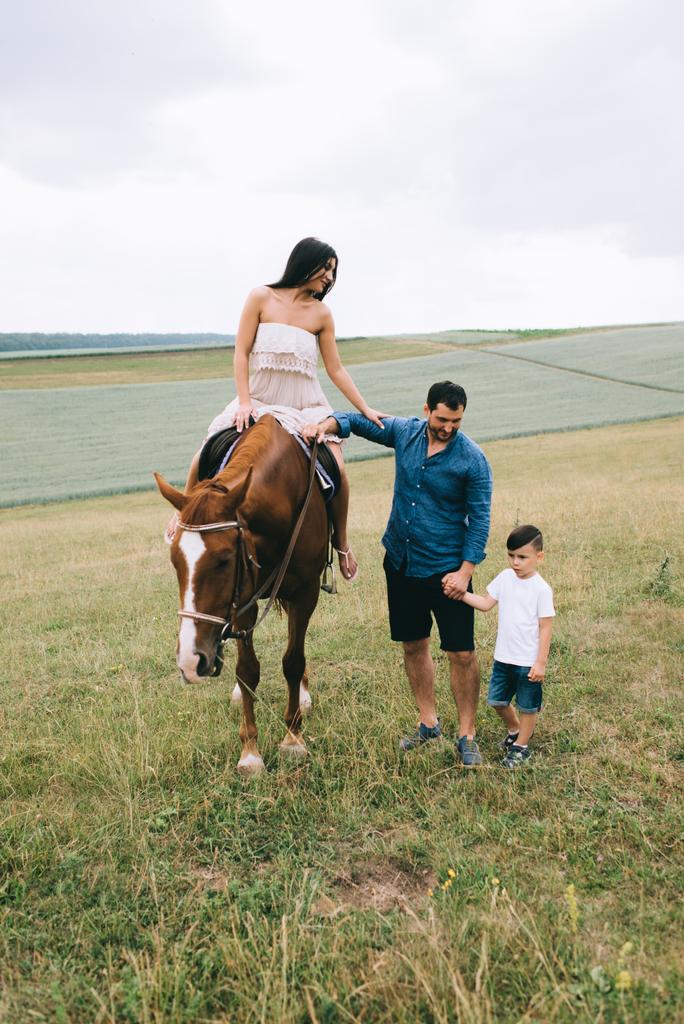 äiti ratsastus ruskea hevonen, isä ja poika seisoo kentällä
 - Valokuva, kuva