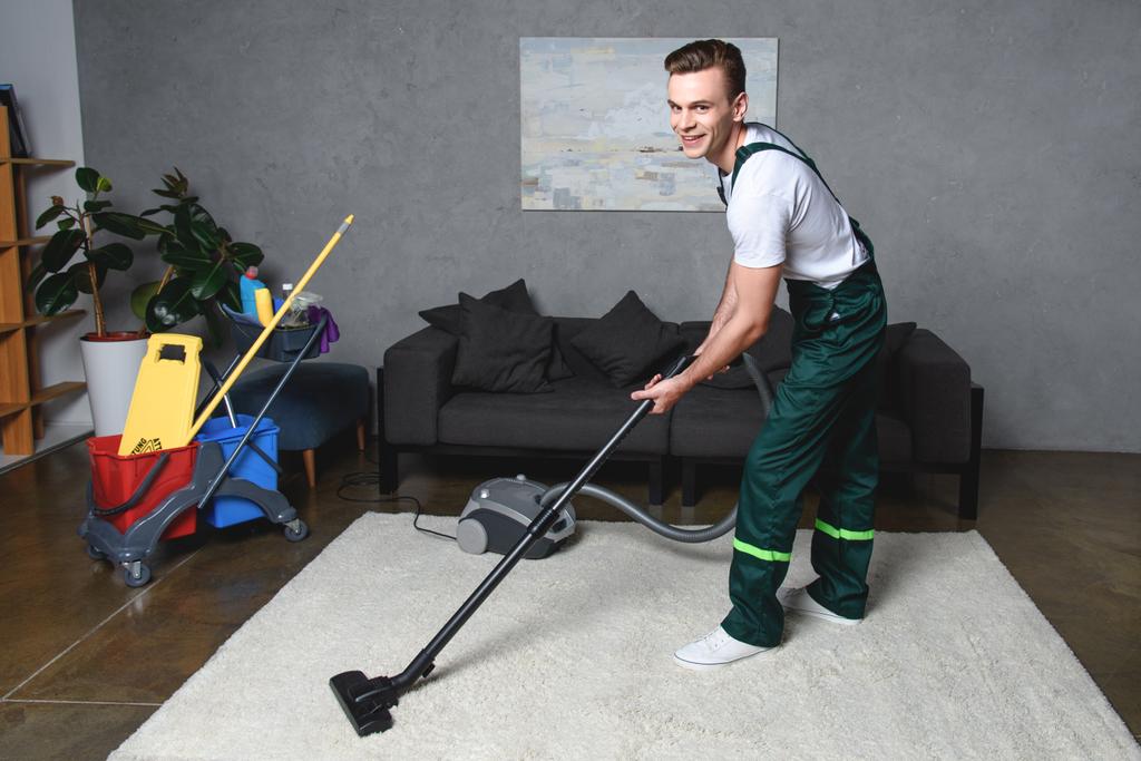 handsome young man cleaning white carpet with vacuum cleaner and smiling at camera - Photo, Image
