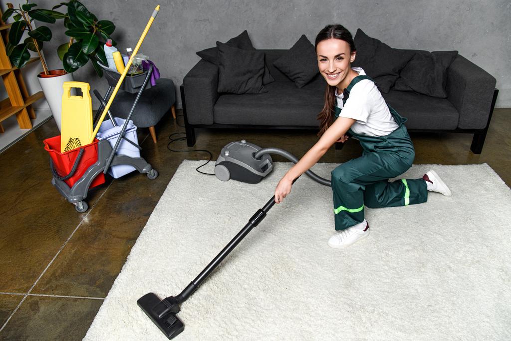 vista ad alto angolo di felice giovane donna pulitore utilizzando aspirapolvere e sorridente alla fotocamera durante la pulizia tappeto bianco
 - Foto, immagini