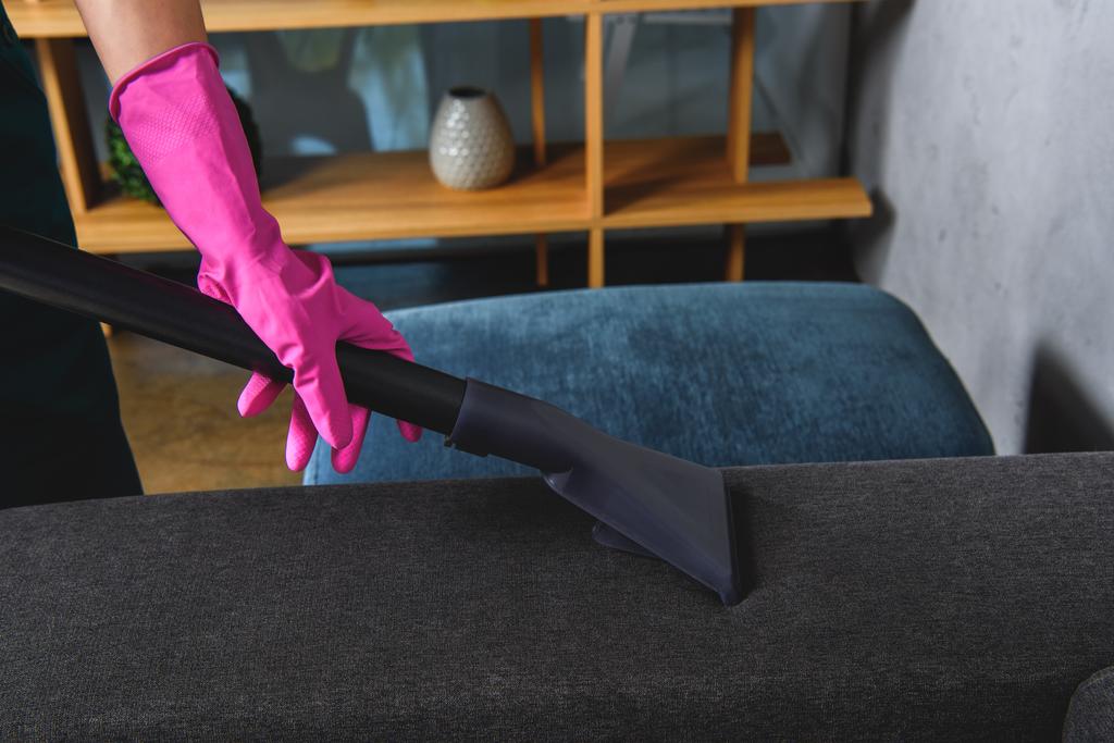 Teilansicht einer Person in Gummihandschuhen, die Möbel mit Staubsauger reinigen   - Foto, Bild