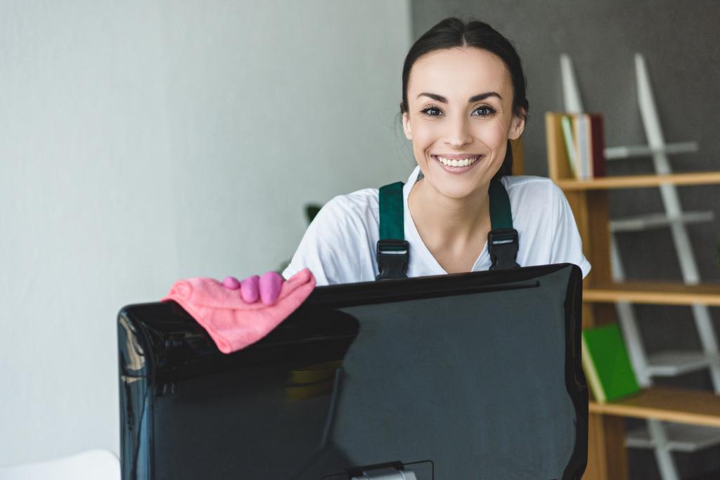 atraente jovem mulher limpeza computador monitor e sorrindo para a câmera
 - Foto, Imagem