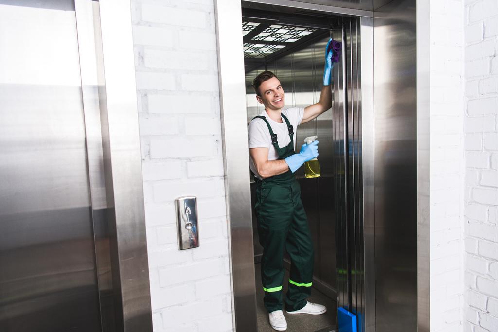 bonito jovem zelador limpeza elevador e sorrindo para a câmera
   - Foto, Imagem