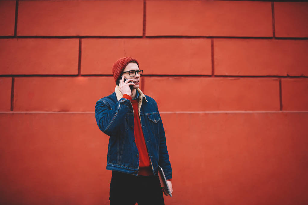 Grave studente hipster vestito in giacca di denim che tiene il computer portatile in mano mentre comunica sullo smartphone durante la telefonata all'operatore in piedi all'aperto su sfondo promozionale rosso
 - Foto, immagini