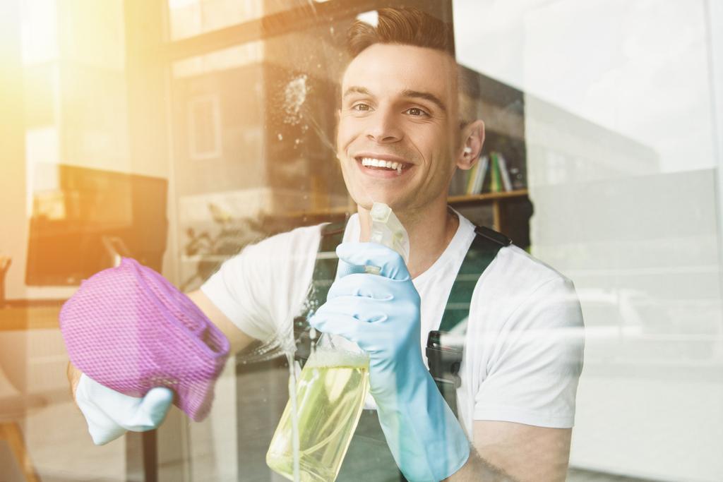 knappe lachende jonge man venster van de schoonmaak en afvegen met sproeiflacon en doek - Foto, afbeelding