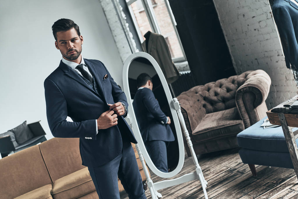 όμορφος άνδρας φαλλοκρατών στο κοστούμι στέκεται στο καναπέ και καθρέφτη   - Φωτογραφία, εικόνα