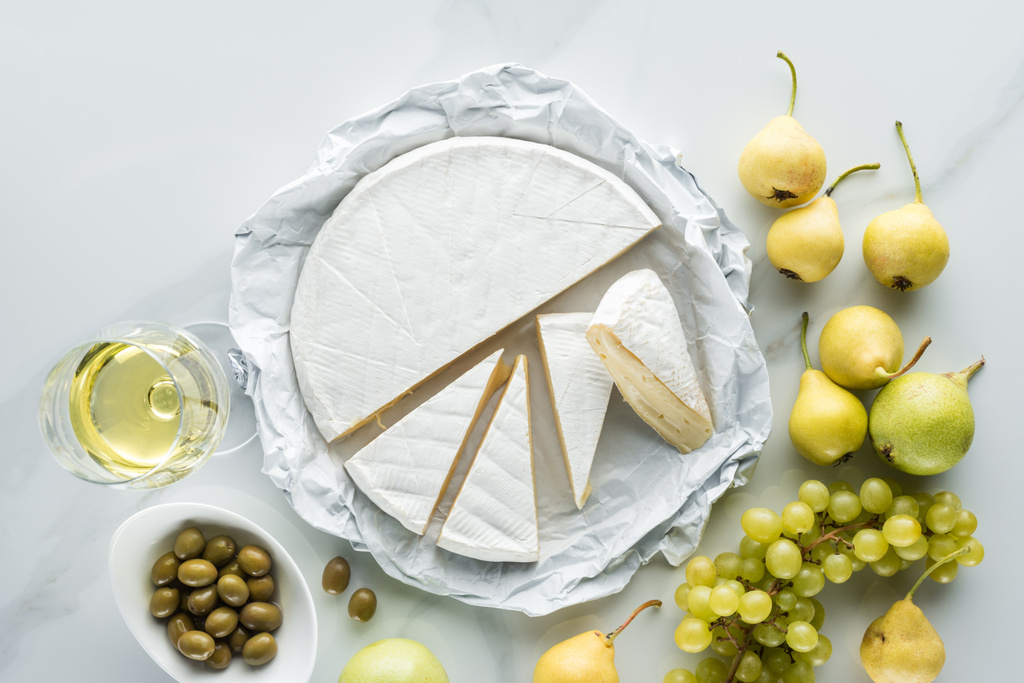 flat lay com copo de vinho, queijo brie, azeitonas e frutas na superfície de mármore branco
 - Foto, Imagem