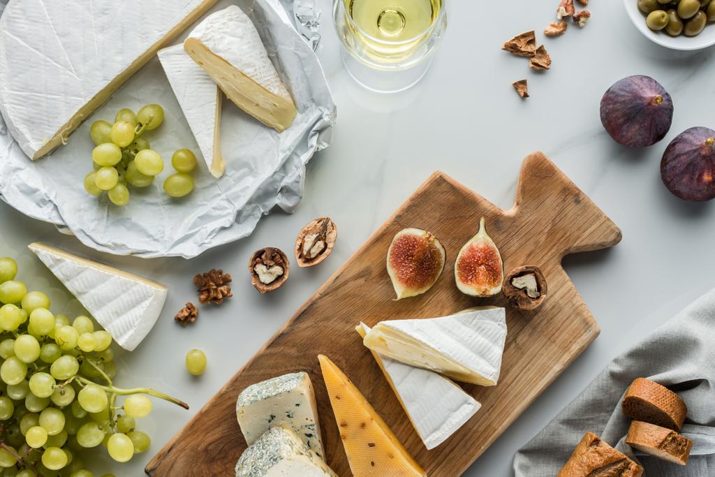 flache Lage mit Essenszusammensetzung aus Käse und Feigenstücken auf Schneidebrett, Obst und Glas Wein auf weißem Marmor - Foto, Bild