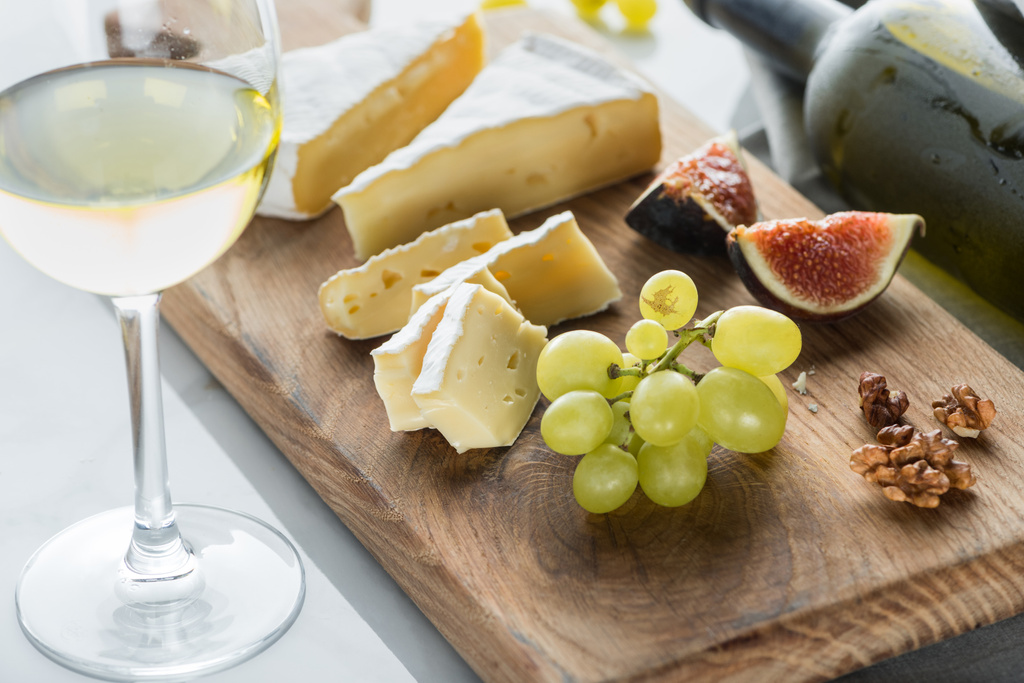 Close-up van camembert kaas, druif en gekonfijte vijgen stukken op snijplank met glas wijn in de buurt door op wit tafelblad - Foto, afbeelding