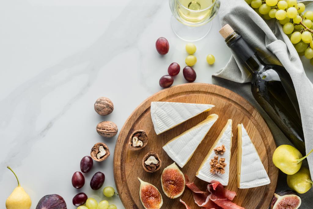 flat lay with wine, cheese, jamon and various fruits arranged on wooden board on white marble tabletop - Photo, Image