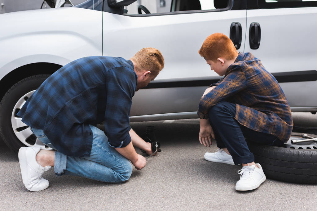 father lifting car with floor jack for changing tire, son sitting on tire - Photo, Image