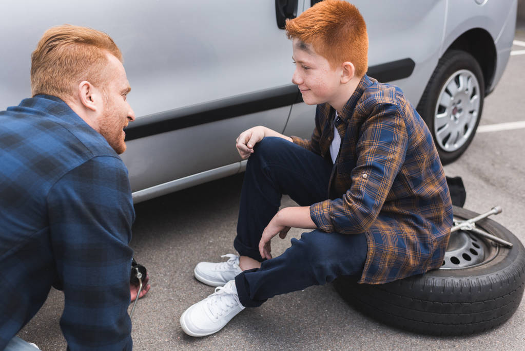 отец и сын меняют шины в машине с валетом и смотрят друг на друга
 - Фото, изображение