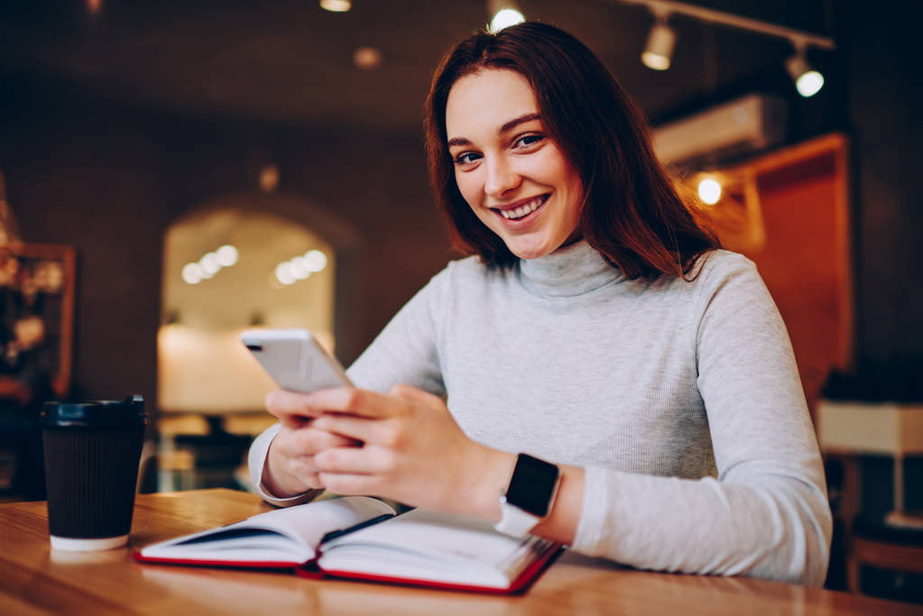Muotokuva iloinen hipster tyttö, joka käyttää älypuhelinta juttelemiseen hauskaa lisäämällä ystäviä sosiaalisissa verkostoissa kahvitauolla, onnellinen nuori nainen katselee kameraa, jolla on matkapuhelin yhteydessä wifiin
 - Valokuva, kuva