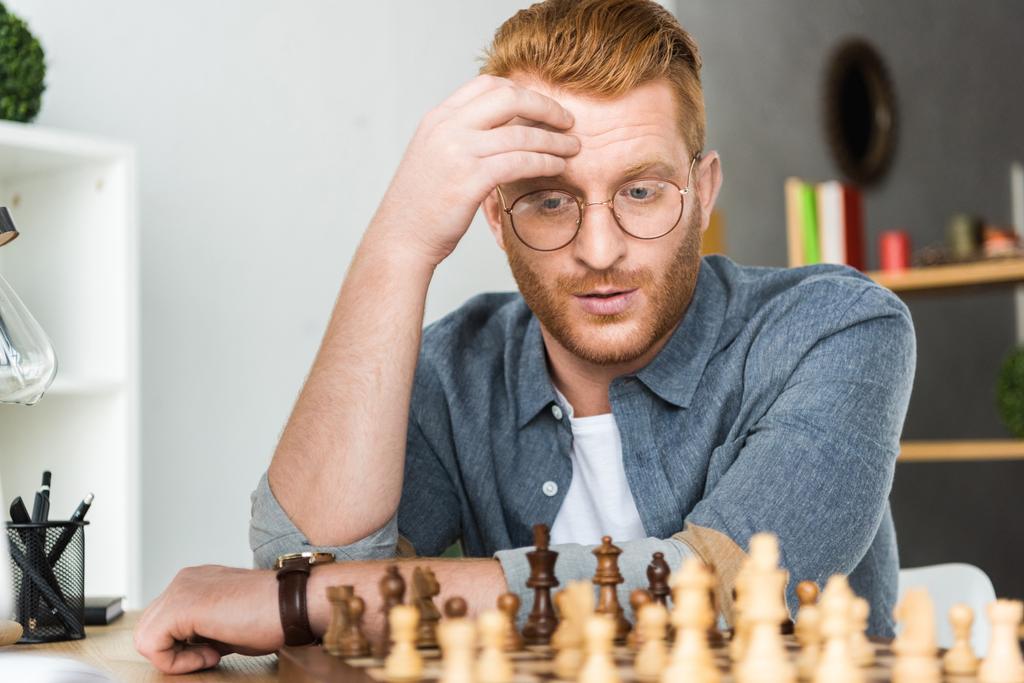 bonito pensivo cabelo vermelho homem olhando para tabuleiro de xadrez em casa
 - Foto, Imagem