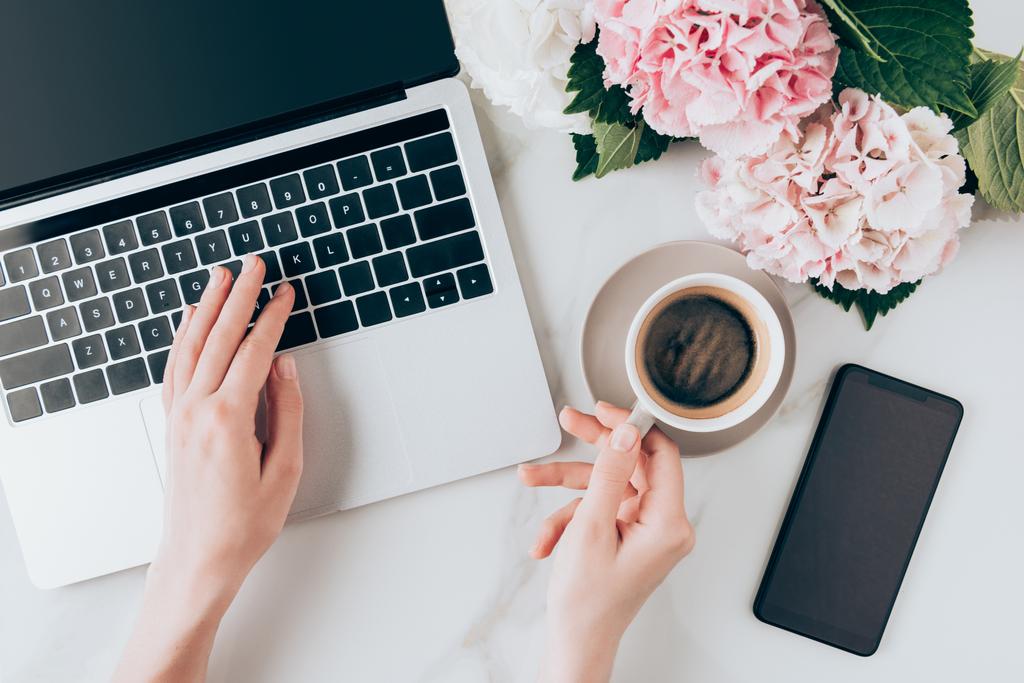 oříznutý pohled ženy pomocí notebooku a hospodářství šálek kávy na stole s smartphone a hortensia květy  - Fotografie, Obrázek