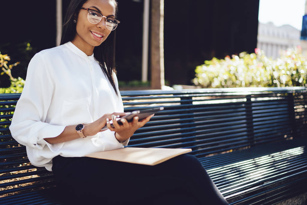 Portrait of positive dark skinned female employee dressed in stylish white shirt and chatting online on digital touch pad resting on bench in city.Successful young woman making transaction on tablet - Photo, Image