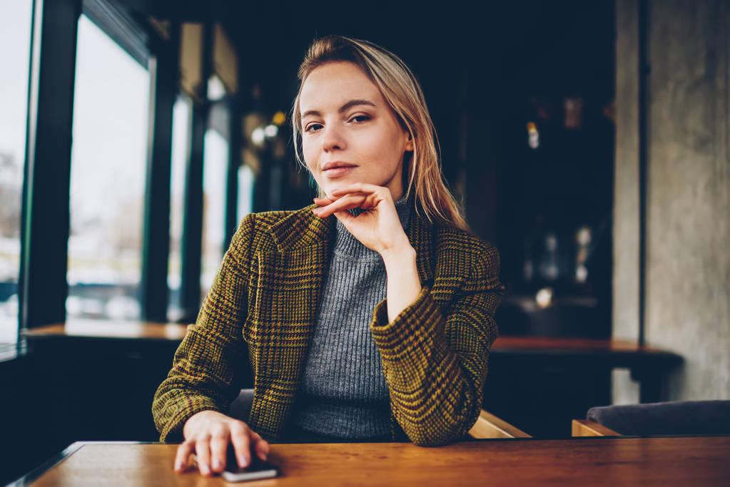Halblanges Porträt einer selbstbewussten Geschäftsfrau in stylischer Jacke, die in die Kamera blickt. Ernsthafte Unternehmerin mit Smartphone in der Hand sitzt an einem Holztisch im Café-Inneren - Foto, Bild