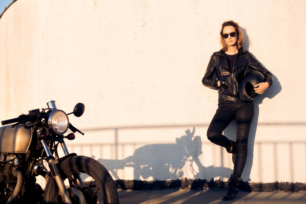 Sexy biker vrouw in zwarte lederen jas en zonnebril met vintage aangepaste caferacer motorfiets in de buurt van de muur. Stedelijke dak parkeren, zonsondergang in de grote stad. Reizen en actieve levensstijl van de hipster. Meisjes macht. - Foto, afbeelding
