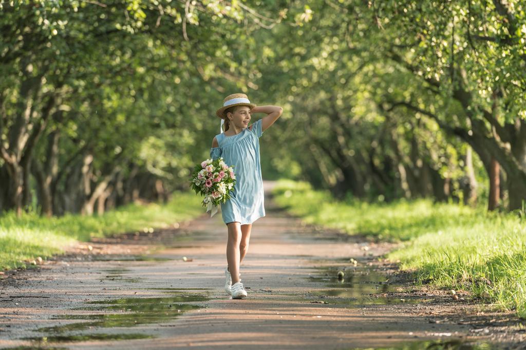 schöne weibliche Kind in trendigem Kleid und Strohhut hält Blumenstrauß und geht in grünen Obstgarten - Foto, Bild