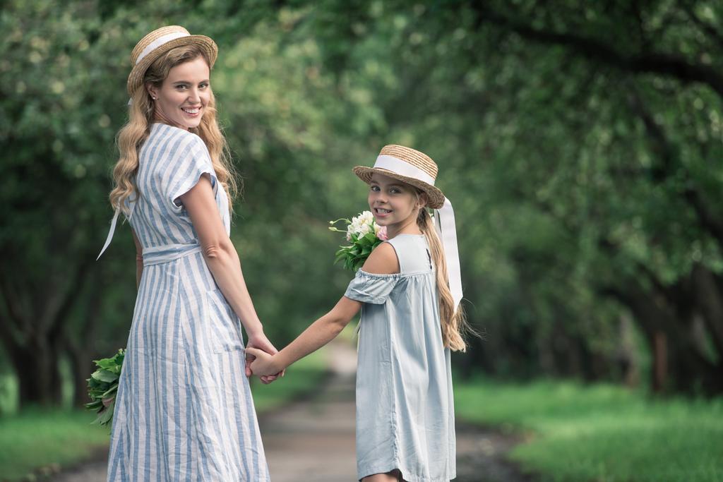 mother and daughter in straw hats holding hands and walking in green park - Photo, Image