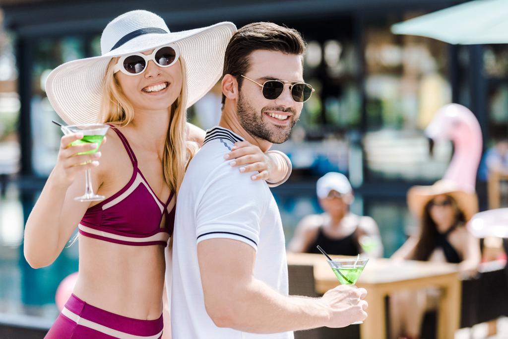 casal feliz em óculos de sol posando com copos de coquetéis perto da piscina
  - Foto, Imagem