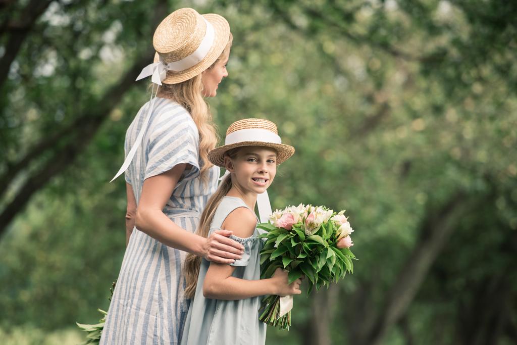 mom and daughter in straw hats with flower bouquets walking in green park - Photo, Image
