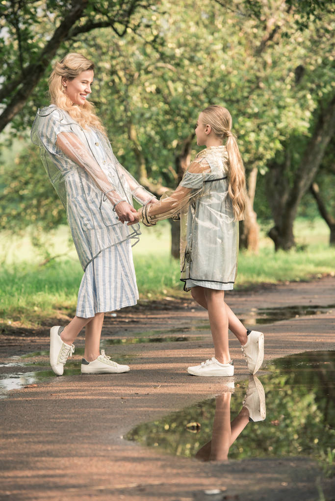 atrakcyjne matka i córka Blondynka w przezroczysta płaszcze, trzymając się za ręce na mokrej drodze w parku - Zdjęcie, obraz
