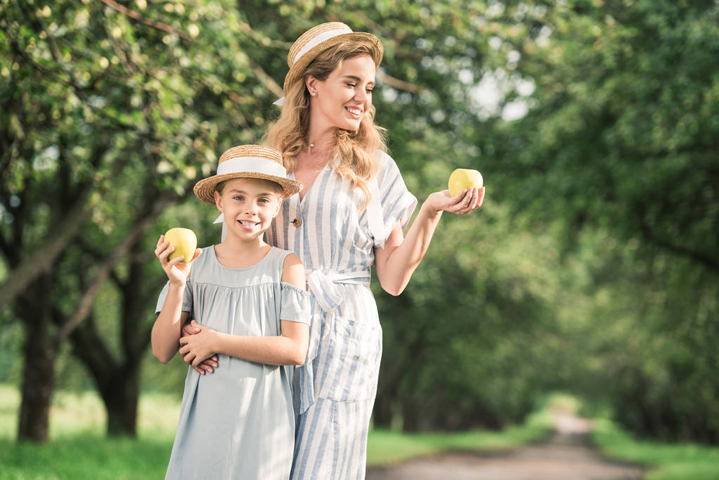 Lächelnde Mutter und Tochter mit Strohhüten, die Äpfel in den Händen betrachten - Foto, Bild