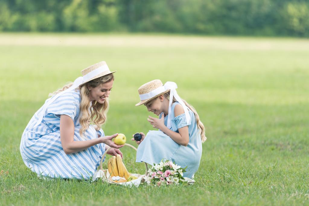 beautiful mother and happy daughter having picnic with fruits and bouquet on green lawn - Photo, Image