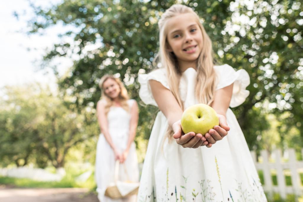 preteen filha segurando maçã, jovem mãe com cesta de vime no fundo, foco seletivo
 - Foto, Imagem