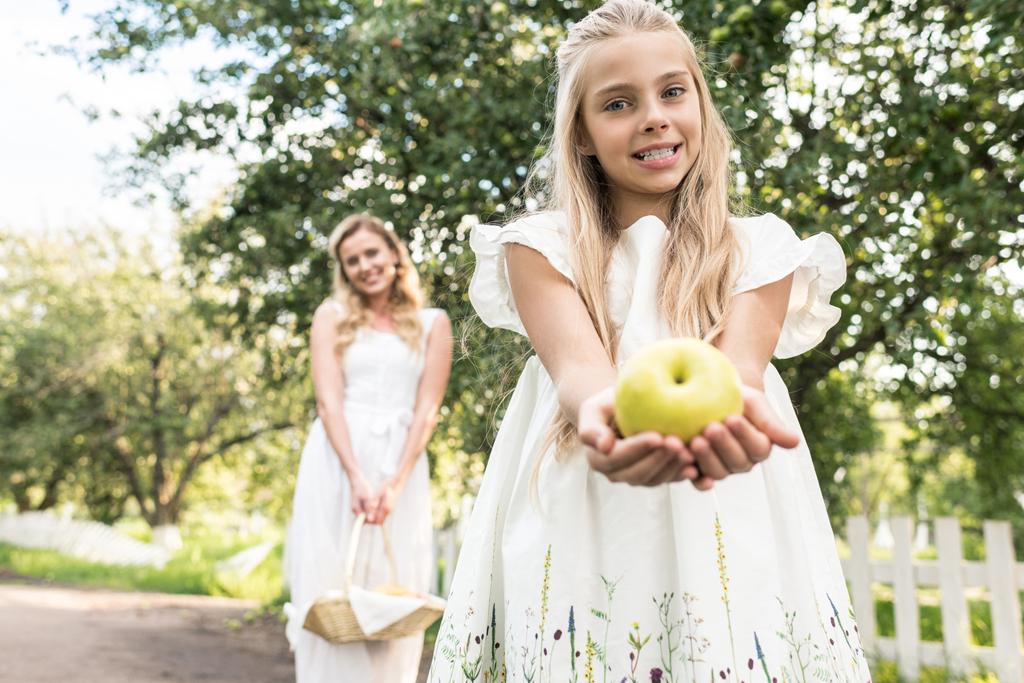 очаровательная дочь держит яблоко, мать с плетеной корзиной на заднем плане
 - Фото, изображение