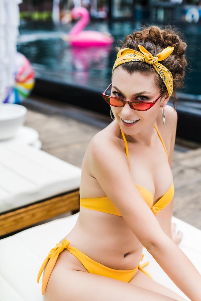 viehättävä nuori nainen keltaisessa uimapuvussa katselee kameraa uima-altaan lähellä
  - Valokuva, kuva
