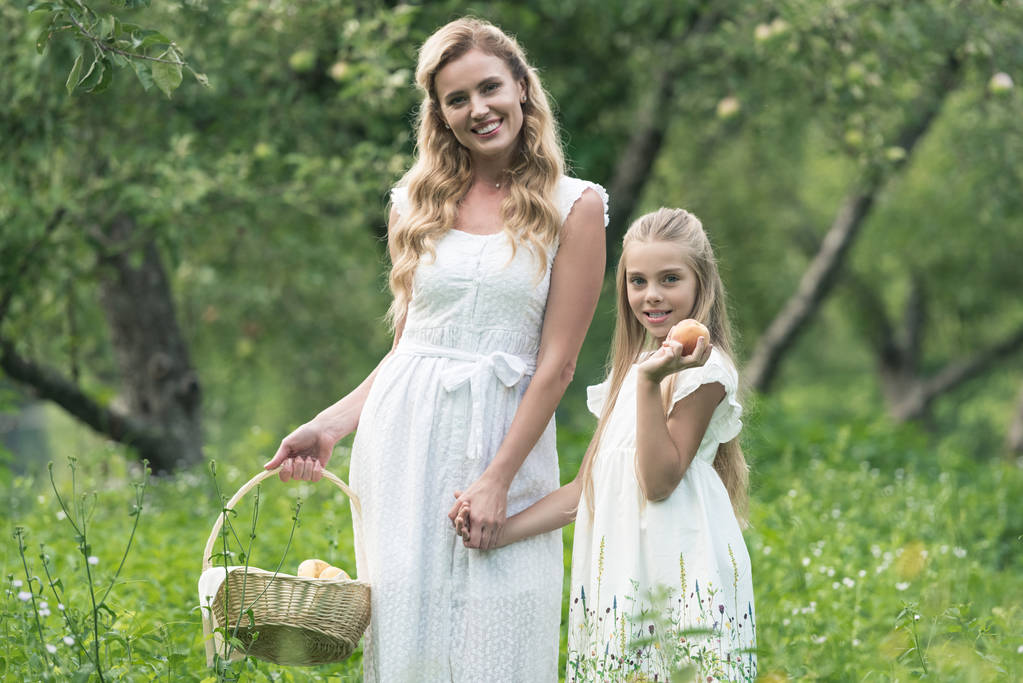 beautiful mother and daughter holding wicker basket with fruits in orchard - Photo, Image