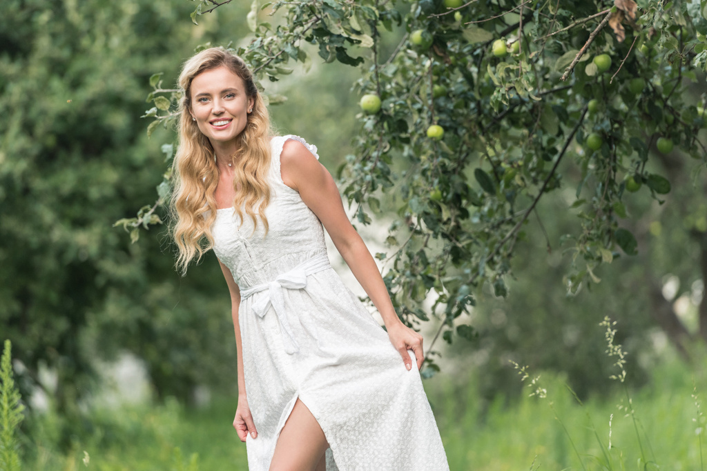 όμορφη χαμογελαστή γυναίκα στην Λευκή φόρεμα, θέτοντας σε περιβόλι με τα δέντρα μηλιάς - Φωτογραφία, εικόνα