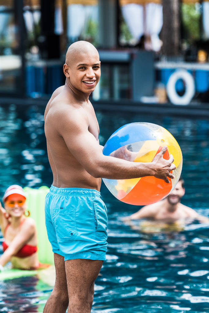 junger muskulöser Mann mit Schwimmball blickt in die Kamera, während seine Freunde  - Foto, Bild