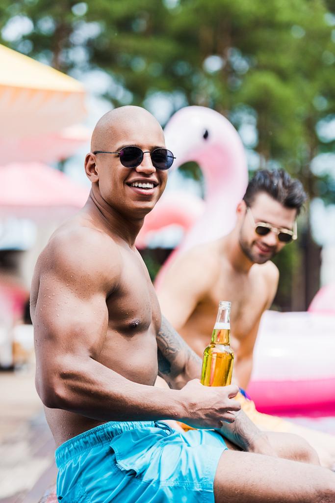 lächelnder muskulöser Mann mit Bier, der in die Kamera blickt, während sein Freund am Pool hinter ihm sitzt  - Foto, Bild