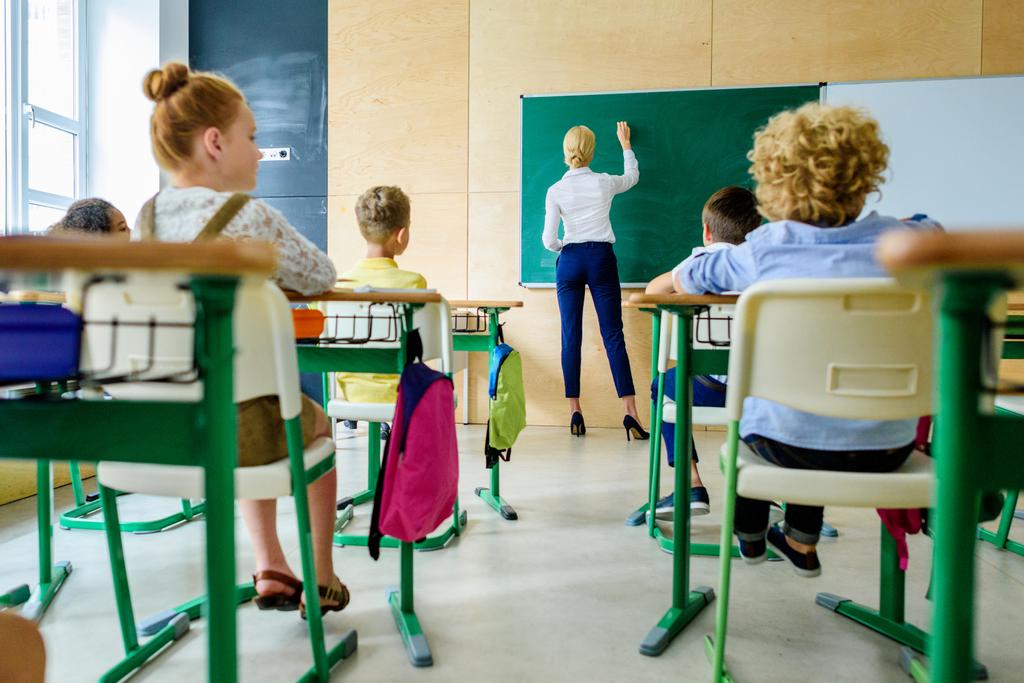 vista trasera de los escolares mirando a la maestra mientras escribe en pizarra
 - Foto, Imagen