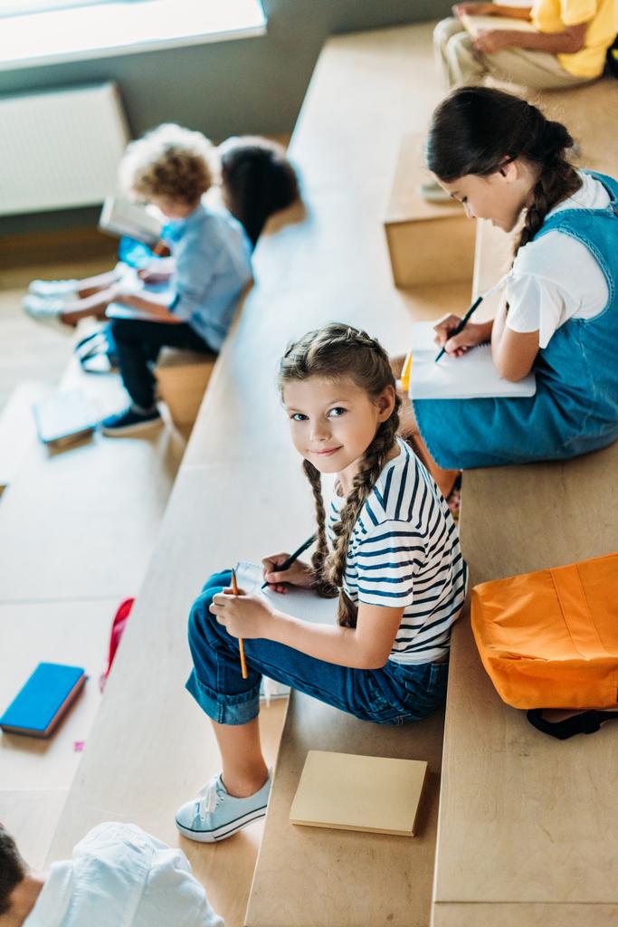 high angle view of schoolgirl with notebook studying on tribune at school corridor while her classmates sitting on background - Photo, Image